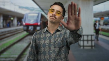 Gözlüklü genç adam dışarıda, tren istasyonunda hareketsiz duruyor. Arka planda kırmızı bir tren var. Seyahat ve kentsel yaşam tarzını öneriyor..
