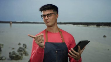 Deniz kenarında tablet tutan önlük giyen bir adam merak gösteriyor. Sahilde düşünceli bir ifade ve arka planda kayalar var..