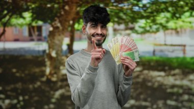 Parkta elinde Norveç banknotlarıyla gülümseyen bir adam çevrenin yeşillikle çevrili olduğunu gösteren olumlu bir hareket sergiliyor..