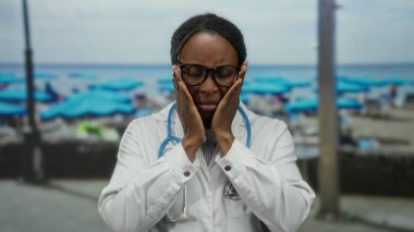 Gözlüklü kadın doktor plajda steteskoplu beyaz bir ceket giyerek güneşli gökyüzünün altında stresli duruyor..