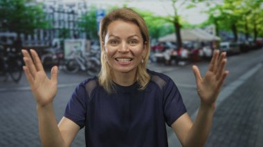 Genç sarışın kadın, bisiklet ve kafe masaları arasında hareketli bir şehir caddesinde açık bir jest ile iki elini kaldırır;.