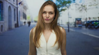 Kadın şehir caddesinde dudaklarını büzüyor. Beyaz bluzlu ve uzun kahverengi saçlı. Gün ışığında yüzünü çerçeveliyor..