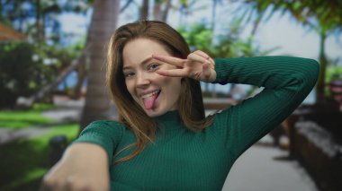 Gülümseyen ve öne doğru eğilen bir kadın yeşil parkta işaret parmaklarını bulanık palmiye ağaçları ile birlikte gösteriyor; neşeli enerji motivasyonu.