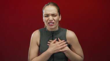 Kırmızı stüdyoda, gergin duruş ve atletik yelek giymiş genç beyaz kadın debriyaj göğsü. Acı kalp krizi..