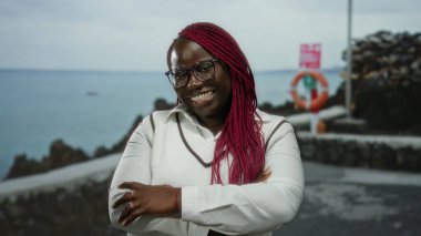 Bir kumsalda çapraz kollarıyla güvenle gülümseyen bir kadın bulutlu bir gökyüzünün altında deniz manzaralı, gözlüklü ve beyaz bir süveter giyen, açık hava manzaralı deniz manzaralı bir tatil atmosferi öneriyor..