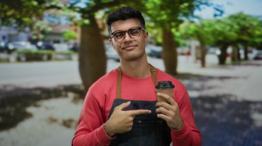 Şehir caddesinde elinde kahve fincanı tutan kırmızı gömlekli ve kot önlüklü genç adam rahat bir şehir manzarası yaratıyor..