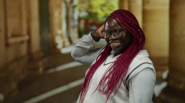 Uzun örgü ve gözlüklü, beyaz bir kazak giyen ve şehir çevresiyle samimi bir şekilde iç içe olan Afro-Amerikalı bir kadın..