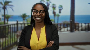 Deniz kenarında, güneşli açık havada palmiye ağaçlarıyla güvenle gülümseyen gözlüklü ve sarı gömlekli bir kadın..