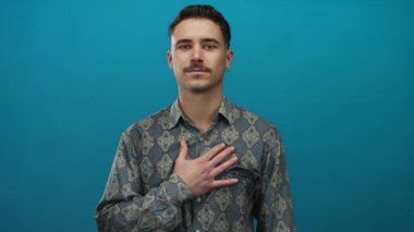 Hispanik genç adam mavi duvara karşı duruyor, kalbi var, duygularını ifade ediyor ve desenli gömlek giyiyor, soyutlanmış arka plan samimiyet konsepti için mükemmel..