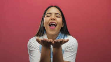 Genç bir kadın, canlı kırmızı bir arka plana gülümserken öpücükler savuruyor, sıcaklık ve neşe taşıyor..