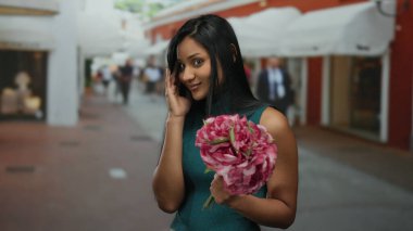 Çiçekleri olan genç bir kadın, arka planda insanlar ve dükkanlarla dolu canlı bir sokakta dalgın dalgın dalgın bakıyor, bir zarafet dokunuşuyla şehir ortamını yakalıyor..