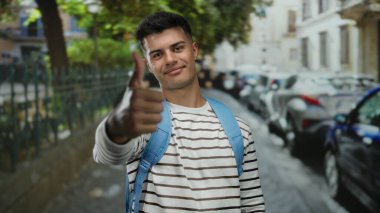 Şehir caddesinde başparmağıyla gülümseyen genç bir adam, çizgili bir gömlek ve mavi bir sırt çantası, park etmiş arabalar ve ağaçlarla çerçevelenmiş, dışarıda pozitiflik yayıyor..