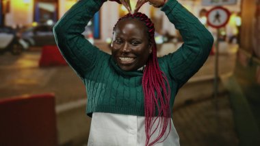 Kollarıyla gülümseyen ve kafasında kalp şekli oluşturan bir kadın. Canlı bir şehir sokak ortamında..