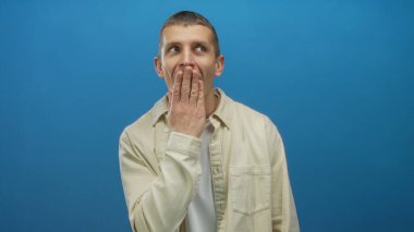 Şaşırmış görünen bir adam, mavi izole edilmiş arka plan karşısında elleri ve ağzını kapatır. Günlük gömlekler giyer, duygularını ve ifadesini ifade eder.