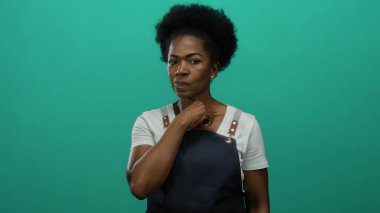 Önlüklü kadın tetikte ve yeşil arka plana karşı tehdit oluşturuyor. İfadeler ve el işaretleriyle yoğun bir odaklanma ile iddialı iletişim kuruyor..