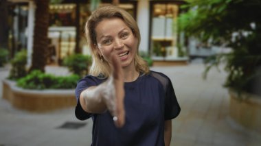 Mavi gömlekli sarışın kadın, gün ışığında gülümserken avluda el sıkışmak için elini uzatıyor; güven güvenilirliği.