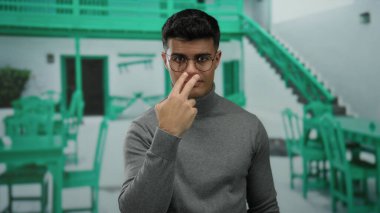 Dışarıda terasta jestler yapan genç bir adam, gözlüklü ve gri kazaklı, yeşil sandalyeli ve masalı bir şehir restoranının arka planında.