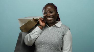 Mavi bir zemin üzerinde alışveriş torbalarıyla gülümseyen bir kadın gözlük takıyor ve başarılı bir alışveriş deneyiminden sonra sevinç ve memnuniyet taşıyan gri bir kazak giyiyor..