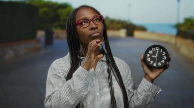 Deniz kenarında saat tutan gözlüklü bir kadın açık hava manzarasının ortasında uzun uzun düşünüyor..