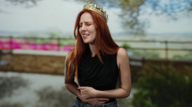 Dışarıda duran kızıl saçlı bir kadın, bir şehir caddesinde rahatsızlığını dile getiriyor, genç ve asil görünümünü şehir ortamında vurguluyor.