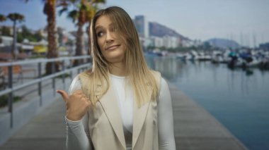 Sarı saçlı genç bir kadın baş parmağıyla bir limanda kayık, güneşli bir sahil ve rahat bir açık hava manzarası sunuyor..