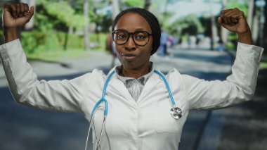 Kendine güvenen doktor kadın şehir ortamında güçlü olduğunu gösteriyor. Neşeli bir ifade takınıyor, gözlük takıyor ve steteskop takıyor. Arka planda canlı yeşillik var.