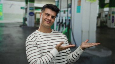 Genç adam bir benzin istasyonunu gösterip gülümsüyor, günlük kıyafetleriyle enerji ve güven taşıyor..