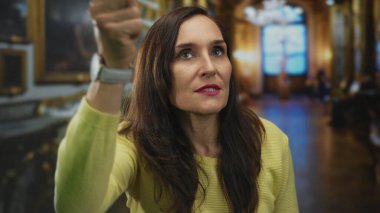 Süslü bir müzede merakını dile getiren bir kadın, evin içinde sarı bir kazak giyiyor, klasik resimlerle çevrili, düşünceli bir müze deneyimini ima ediyor..