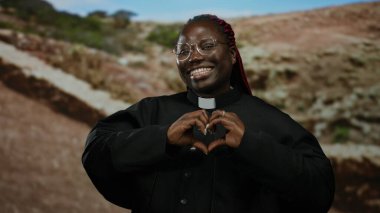 Gözlüklü kadın, kalbi parka yerleştirilmiş ruhban cübbesi giymiş açık arka planı bulanık bir şekilde doğada gülümsüyor.