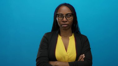 Gözlüklü, kıvırcık saçlı, sarı gömlekli, kolları çapraz siyah ceketli, izole bir mavi duvarın karşısında duran kendine güvenen bir kadın..