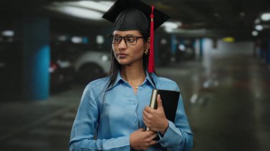 Otoparkta elinde kitap tutan, mezuniyet şapkası ve gözlüğü takan, akademik başarı ve kapalı alanda tefekkür öneren bir kadın..