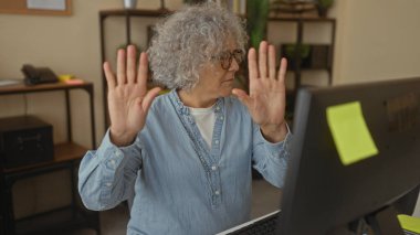Ofis ortamında el kol hareketi yapan, orta yaşlı, kıvırcık saçlı, gözlüklü ve kot gömlekli, bilgisayarın önünde el kaldırarak iletişim hareketi yapan bir kadın..