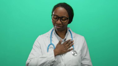 Gözlüklü ve steteskop takan doktor yeşil arka planda tek başına, gözleri kapalı ve elleri göğsünde, sağlık uzmanlarında sakinliği ve çeşitliliği yansıtan bir şekilde duruyor.
