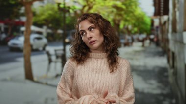 Kazaklı bir kadın ağaçlar ve bulanık bir arka planla sokak ortasında duruyor doğal ışık altında rahat bir şehir atmosferi yaratıyor..