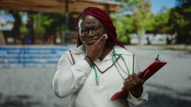 Afrika kökenli Amerikalı bir kadın parkta gözlüklü ve steteskop takmış elinde bir pano, jestlerle ilgili endişelerini dile getiriyor..