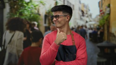 Gözlüklü genç adam, şehir caddesinde hareketli bir baş parmağıyla hareketli hareketler yapıyor. Etrafı bulanık gezginler ve şehir yeşillikleriyle çevrili..