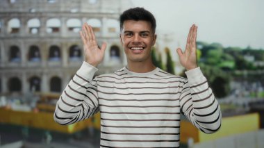 Roma ikonik stadyumunun önünde neşeli bir şekilde poz veren genç adam, açık bir gülümseme ve çizgili bir gömlek sergiliyor, canlı bir şehir atmosferini yakalıyor..