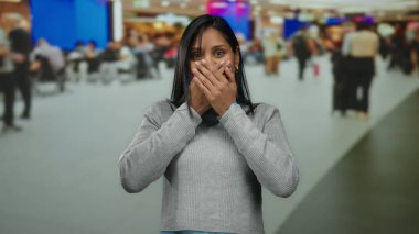 Havaalanı terminalinde şaşırmış bir kadın ellerini kapalı bir şekilde kapalı bir terminalde bulanık gezginler ve parlak işaretlerle canlı bir atmosfer yaratarak ağzını kapatıyor..