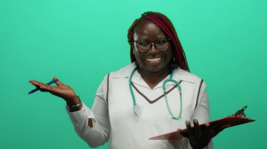 Steteskoplu Afrika kökenli Amerikalı kadın doktor gülümsüyor ve elinde yeşil bir arkaplan, profesyonelliği ve tıbbi kıyafete olan güveni aktarıyor..