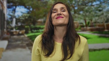 Hispanik kadın canlı yeşil bir açık hava parkında eğleniyor, neşe ve bağlantı ifade ediyor, sarı giyiniyor, elleri neşe içinde gülümsüyor..