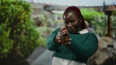 Saçları örülmüş yeşil kazaklı bir kadın açık hava parkında bir parmak tabancasının hareketini taklit ediyor..