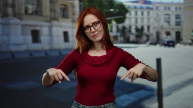 Gözlüklü ve kırmızı kazaklı bir kadın şehir sokaklarında dikiliyor poz veriyor, açık şehir arka planına karşı kızıl saçlarını gösteriyor..