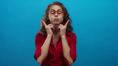 Kıvırcık saçlı ve kırmızı gözlüklü bir kadın canlı mavi bir arka plana karşı neşeli yüzler yapıyor, neşe ve mizah ifade ediyor..