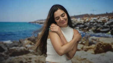 Beyaz atlet giyen genç İspanyol kadın kayalık sahilde çıplak omuzlarına sarılıyor..