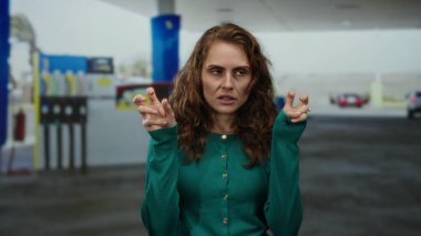 Benzin istasyonunda el kol hareketi yapan bir kadın arka planda bulanık yakıt pompalarıyla açık havada duygularını ve gündelik tarzını sergiliyor..