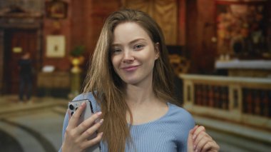 Kadın görünür bir yüzle gülümser ve kilisede ahşap sunakta akıllı telefonu tutar; dinginlik yansıması.