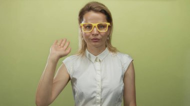 Stüdyoda sarı gözlüklü ve beyaz kolsuz gömlek giyen sarışın kadın el sallıyor;.