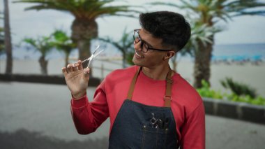 Gözlüklü genç adam elinde makasla, önlükle gülümsüyor, deniz kenarında palmiye ağaçlarıyla dikiliyor, neşeli bir berber teması yaratıyor..