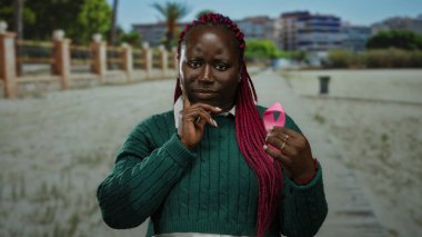 Yeşil kazaklı kadın parkta pembe kurdele tutuyor. Arka planda binalar var. Açık hava ortamında meme kanseri farkındalığı ve desteğine odaklanıyor..