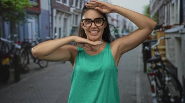Yeşil kollu tişörtlü genç İspanyol kadın gözlüklü çerçeve takıyor ve şehir sokaklarında kollarını kaldırıyor; mutluluk..